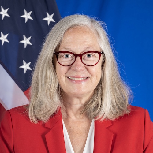 DCM Konick wearing a red jacket standing in front of the U.S. flag